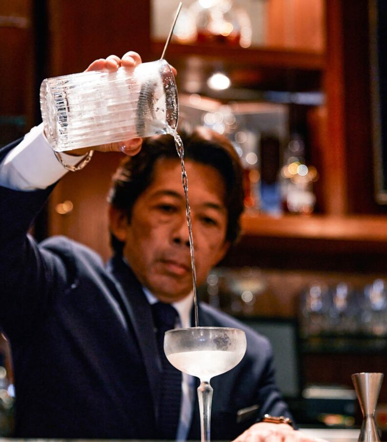 Bartender pouring a cocktail through a strainer into a coupe glass behind a bar counter