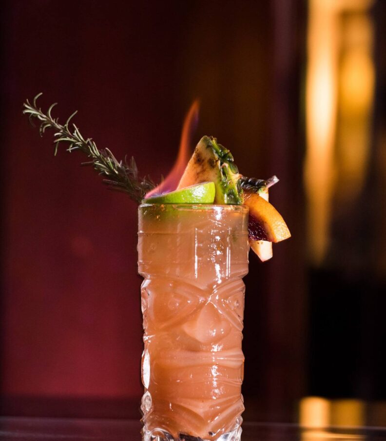 A sculpted tiki-style cocktail rises in warm amber tones, crowned with citrus, rosemary, and a dramatic flicker of flame