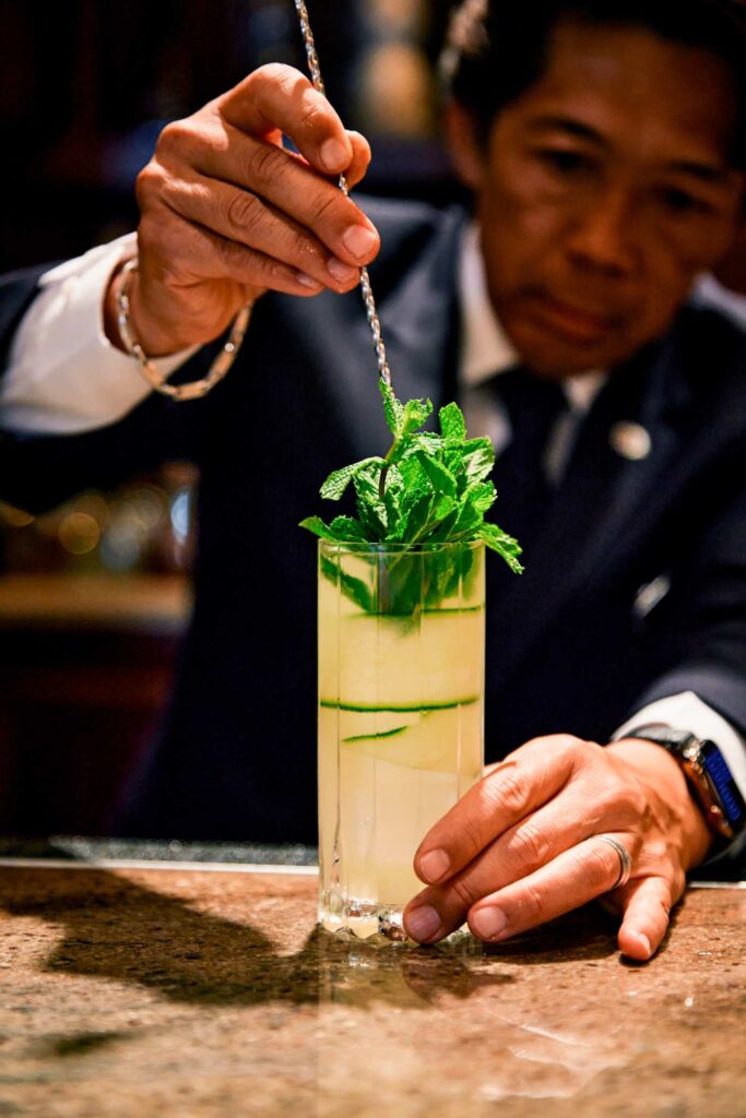 Bartender garnishes a tall pale cocktail with fresh mint using a bar spoon at a marble counter