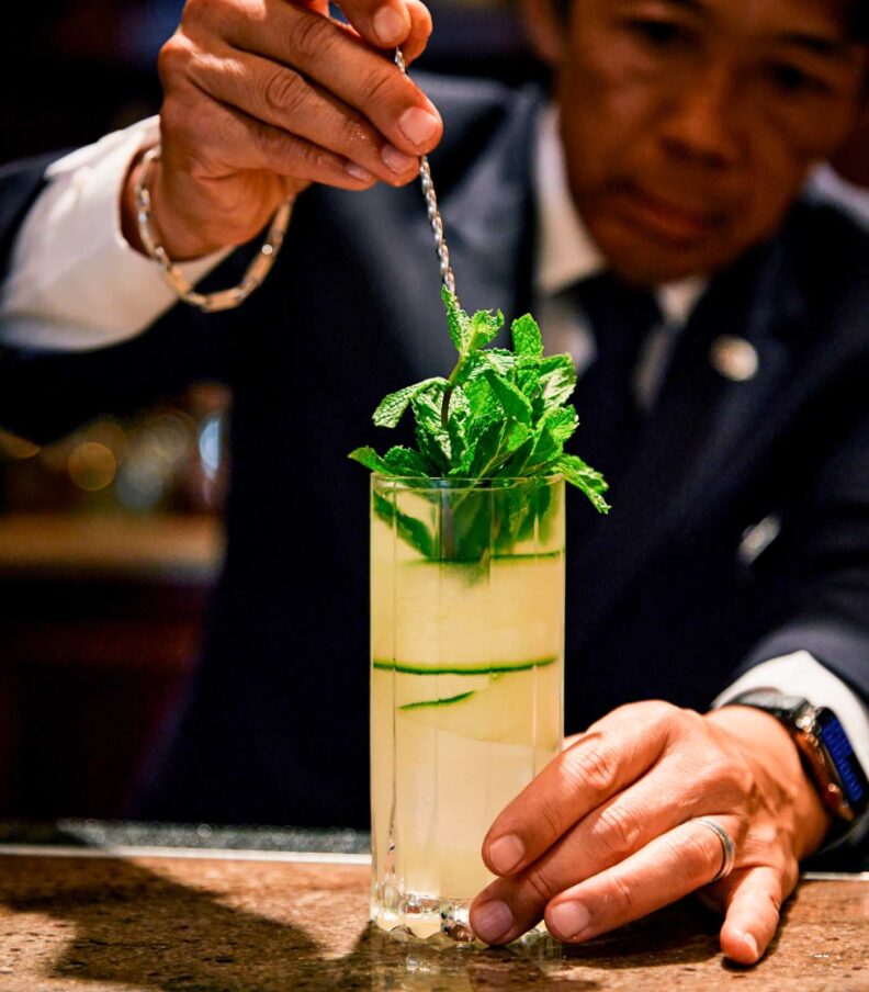Bartender garnishes a tall pale cocktail with fresh mint using a bar spoon at a marble counter