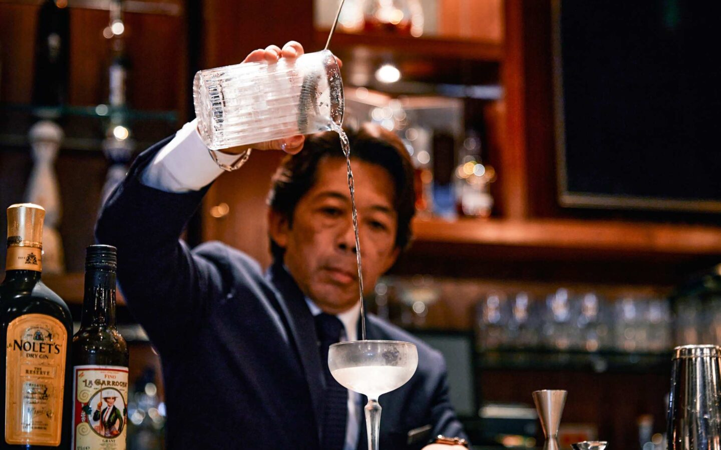 Bartender pours a clear cocktail into a coupe glass behind a polished bar with bottles and glassware in the background