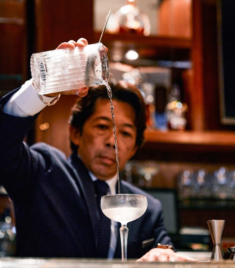 Bartender pours a clear cocktail into a coupe glass behind a polished bar with bottles and glassware in the background