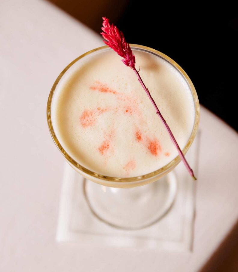 Top view of a creamy cocktail in a coupe glass, garnished with a red floral stem on a white table