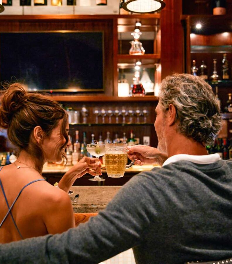 Two people seated at a bar raising glasses in a toast, with shelves of liquor bottles and warm wood paneling in the background