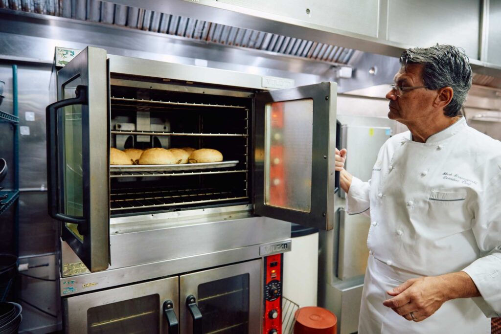Chef in a white jacket checking loaves of bread baking in a commercial oven in a restaurant kitchen