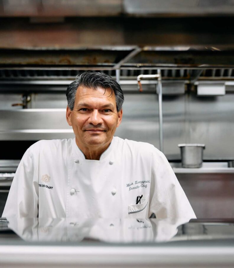 Chef in a white jacket standing behind a stainless steel counter in a professional restaurant kitchen
