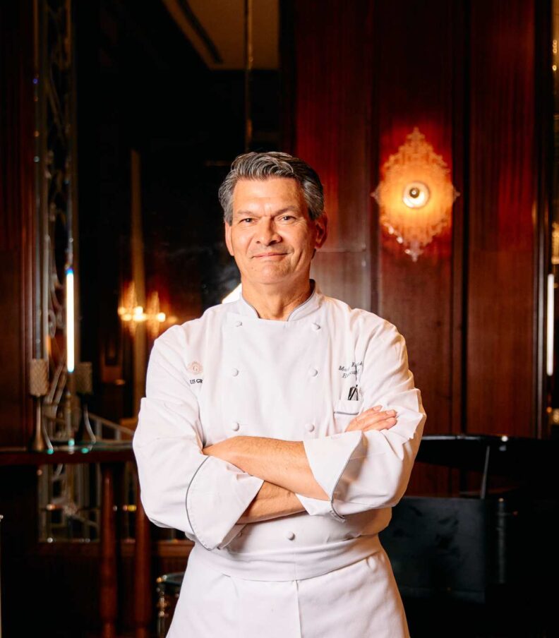 Executive chef standing with arms crossed in a formal restaurant interior wearing a white chef jacket