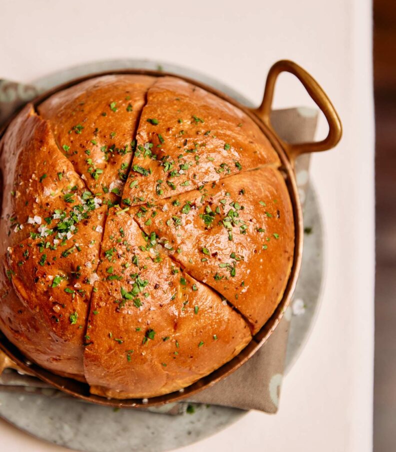 Warm round bread loaf sliced and served in a copper pan with herbs on a white tablecloth