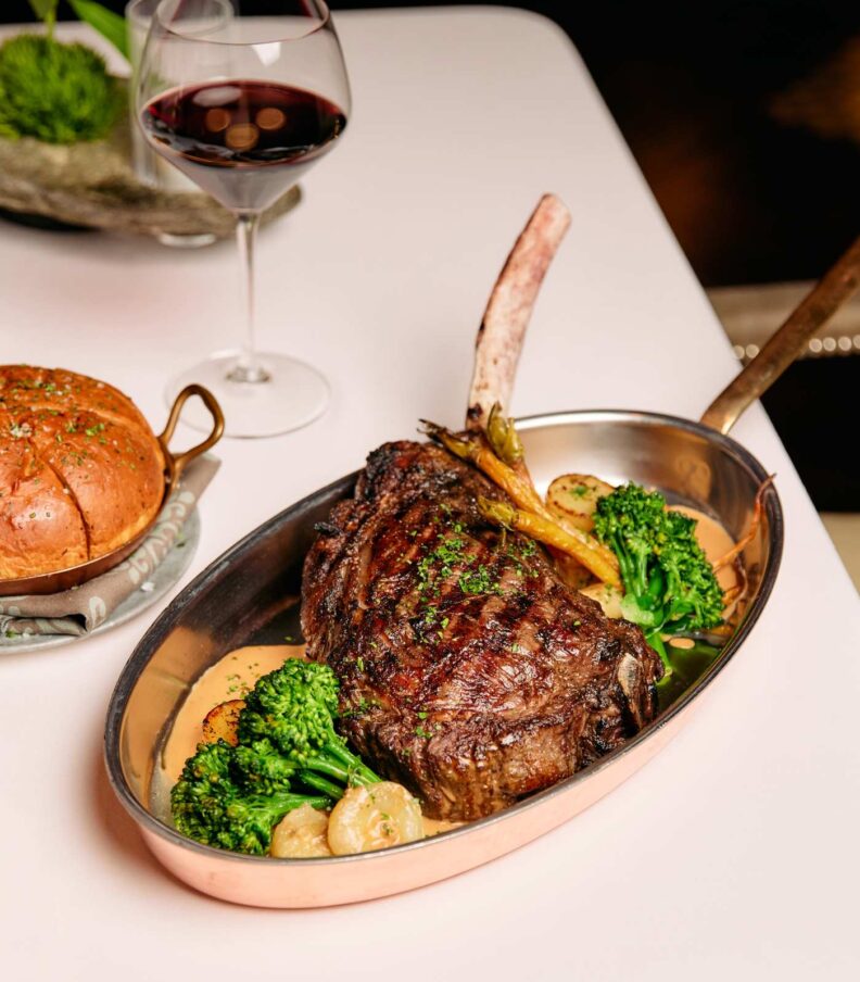 Bone in ribeye steak served in a copper pan with vegetables, bread, and a glass of red wine on a white tablecloth