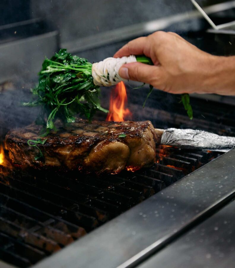 Chef grilling a thick cut steak over open flames while basting with fresh herbs in a professional kitchen