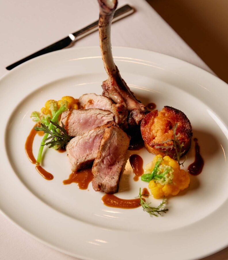 Plated rack of lamb with sliced meat, vegetables, sauce, and a crisp potato element on a white plate
