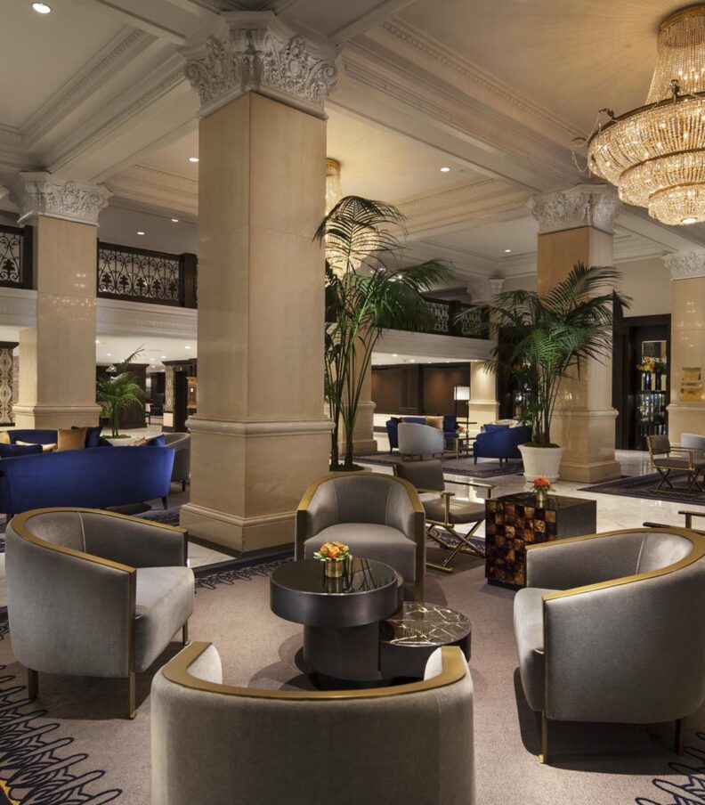 Spacious hotel lobby with chandeliers, marble floors, lounge seating, tall columns, and indoor palm plants