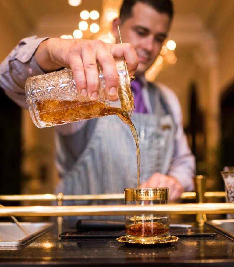 A bartender mid-pour, decanting an amber cocktail from a cut-crystal mixing vessel into a gilded rocks glass. Warm chandelier light, brass rail details, and focused hands convey precision, craft, and classic cocktail theater in an elegant bar setting.