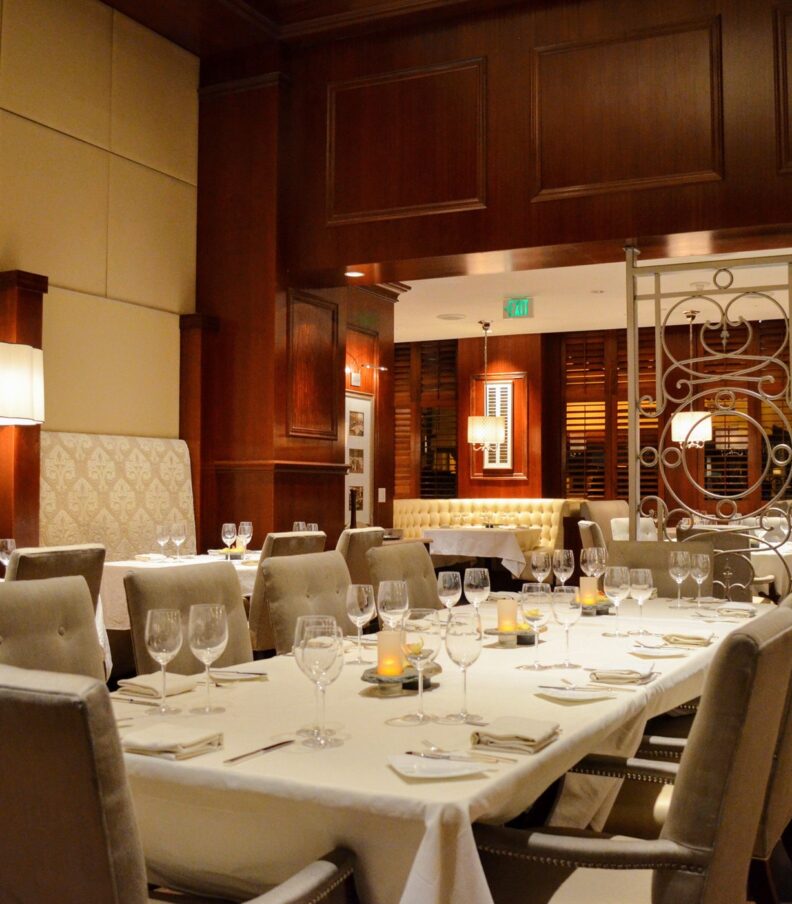Elegant dining room with set tables, upholstered chairs, warm wood paneling, and soft ambient lighting.