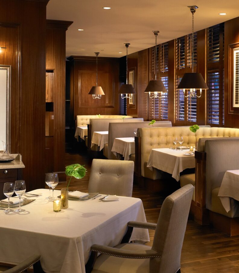 Warm wood paneled dining room with upholstered booths, white tablecloths, and softly lit tables set for fine dining.