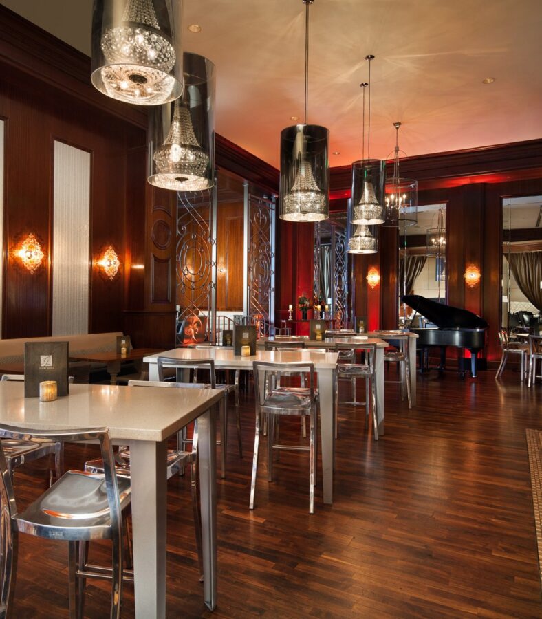 Elegant restaurant interior with polished wood floors, modern tables, bar seating, pendant lights, and a grand piano in the background.