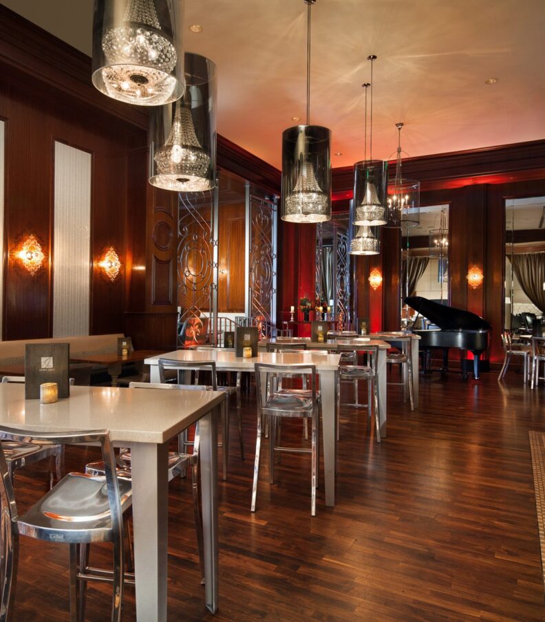 Elegant restaurant interior with polished wood floors, modern tables, bar seating, pendant lights, and a grand piano in the background.