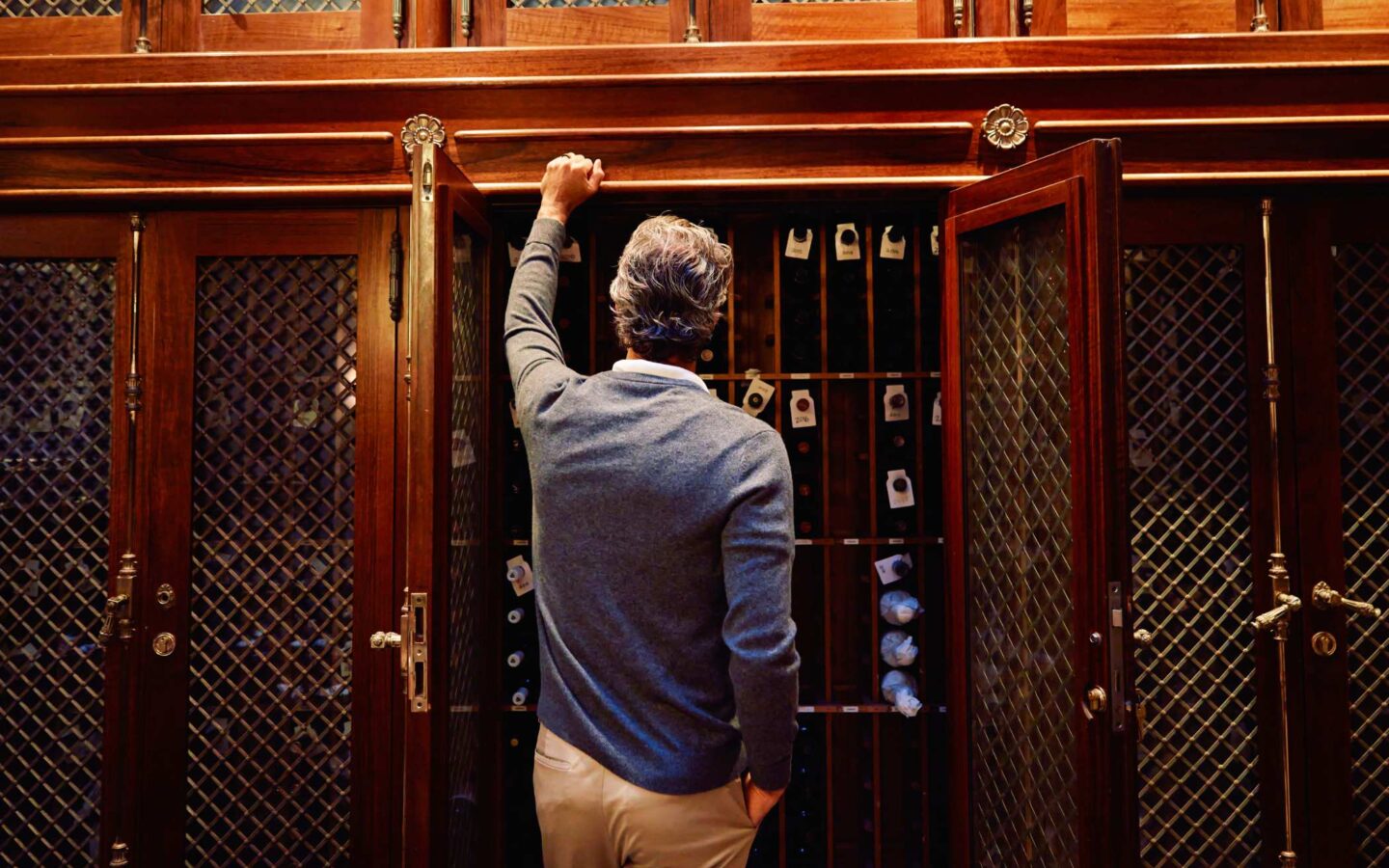Person selecting a bottle from a wooden wine cabinet with glass lattice doors in a restaurant cellar setting