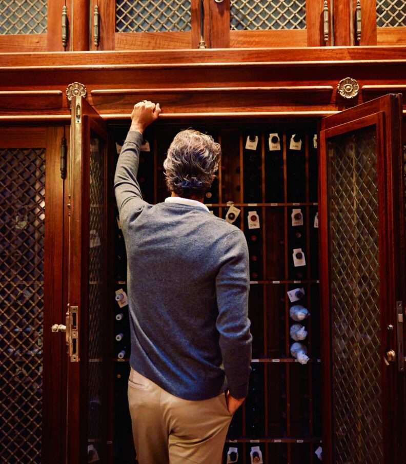 Person selecting a bottle from a wooden wine cabinet with glass lattice doors in a restaurant cellar setting