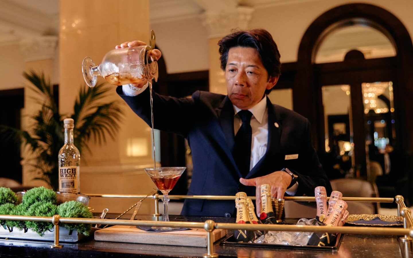 Bartender pouring a cocktail tableside from a glass decanter in an elegant hotel lobby bar setting