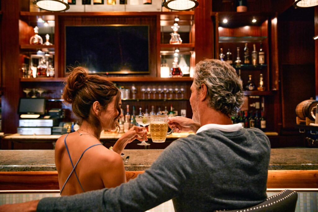 Two people seated at a bar raising glasses in a toast, with shelves of liquor bottles and warm wood paneling in the background