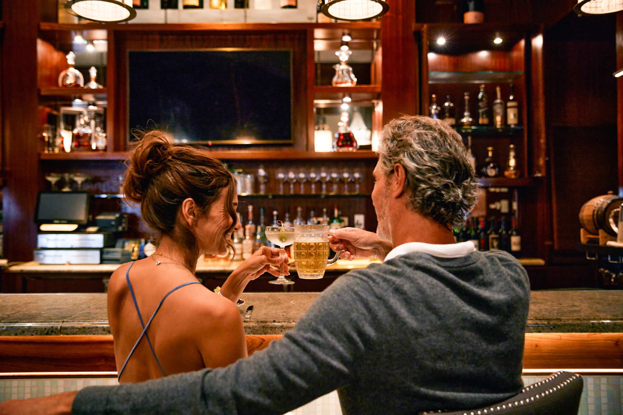 Two people seated at a bar raising glasses in a toast, with shelves of liquor bottles and warm wood paneling in the background