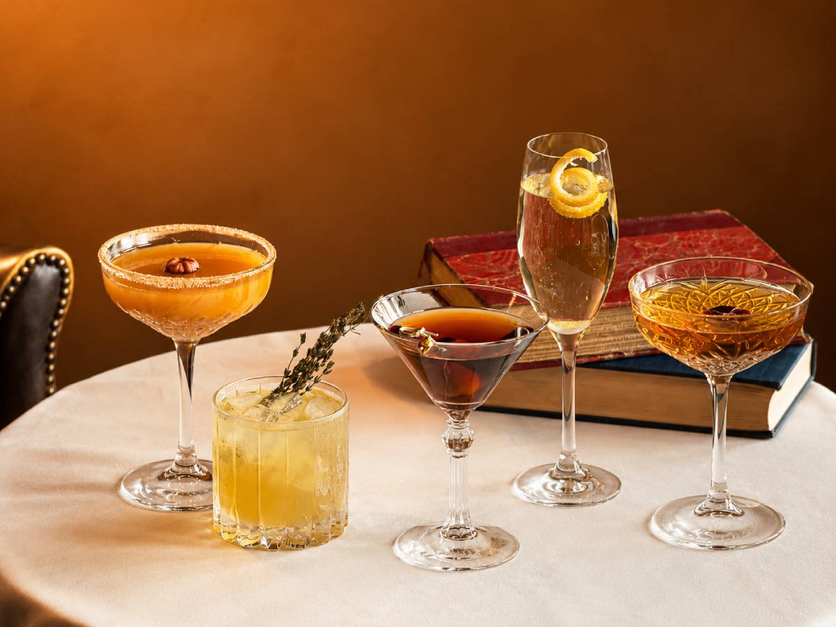 Assorted cocktails in coupe, flute, and rocks glasses arranged on a table with vintage books.
