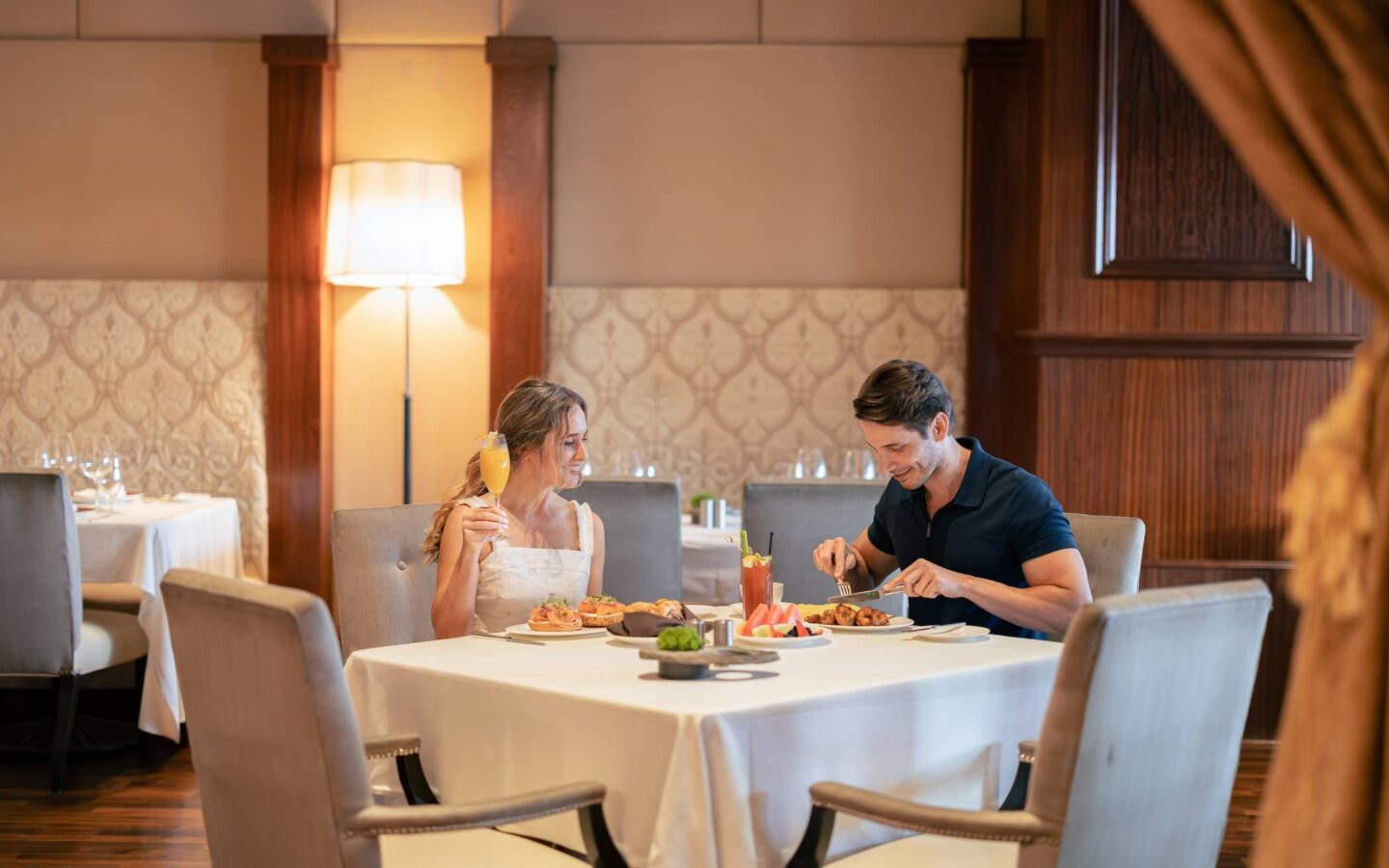 Couple enjoying a meal and cocktails at an elegant restaurant dining room with white tablecloths and warm wood accents.